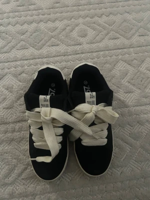 Svarta sneakers från ZigZag - Snygga svarta sneakers från ZigZag med vita detaljer och breda vita skosnören. Skorna har en vit sula med brunt yttersula och en vit rand på sidan. Ovandelen är i textil och mockaimitation. Perfekta för en avslappnad streetstil.
