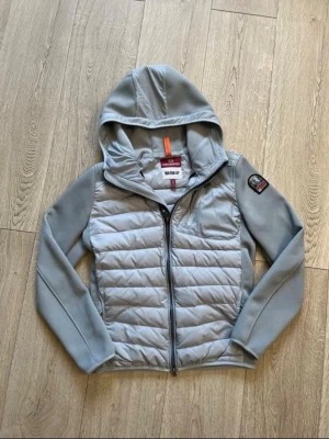 Ljusgrå pufferjacka från Parajumpers - Snygg ljusgrå pufferjacka från Parajumpers med huva och dragkedja framtill. Jackan har quiltad front, släta ärmar och ett tydligt PJS-märke på ärmen. Perfekt för kalla dagar och har en modern, sportig look.