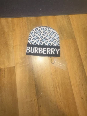 Burberry mössa med logotypmönster - Snygg mössa från Burberry med stort logotypmönster i vitt och ljusblått på svart botten. Tydlig BURBERRY-text framtill och klassisk design. Tillverkad i mjukt stickat material som känns bekvämt och stilrent. Perfekt accessoar för dig som vill sticka ut.