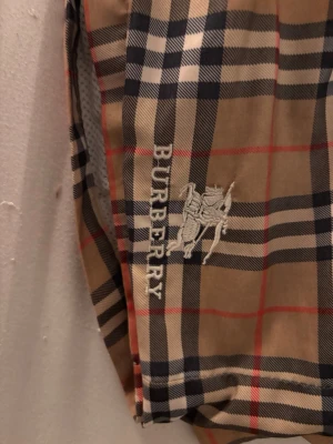 Rutig Burberry shorts med snörning - Snygga shorts från Burberry i klassiskt beige, svart, vit och röd rutig design. Broderad Burberry-logga och riddare på ena benet. Shortsen har elastisk midja och snörning samt meshfoder på insidan. Perfekta för sommaren och streetwear.