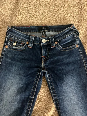 Mörkblå lowwaist jeans från true religion - Snygga mörkblå bootcut jeans från true religion med klassiska fem fickor och orange kontrastsömmar. Lite slitna nere vid benet och en liten fläck vid knät där färgen gått bort.