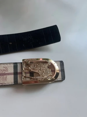 Rutig skärp från Burberry med guldspänne - Säljer ett snyggt skärp från Burberry med klassiskt rutigt mönster i beige, svart och rött. Skärpet har ett exklusivt guldigt spänne med Burberry-logga och är tillverkat i skinn. Perfekt för att lyfta din outfit.