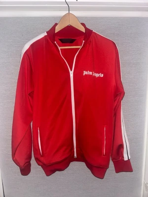 Röd zip hoodie från Palm Angels - Snygg röd zip hoodie från Palm Angels med vita detaljer och logga på bröstet. Klassisk sportig look med vita ränder längs ärmarna och ribbade muddar. Tillverkad i ett glansigt material som ger en retrovibe. Perfekt för dig som gillar streetwear och vill sticka ut.