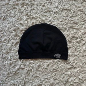 SYNA skullcap - Svart, stilren mössa från SYNA med diskret logga broderad framtill. Tillverkad i mjukt och stretchigt material som sitter skönt på huvudet. Perfekt för dig som gillar enkel och clean stil.