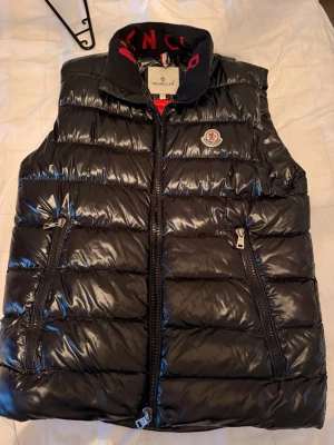 Svart dunväst  - Snygg svart dunväst  med glansig finish, två dragkedjefickor och hög krage med röd logotyptext. Insidan är röd med tryck och serietidningsdetalj. Klassisk Moncler-logga på bröstet och dragkedja framtill. Perfekt för lager på lager och streetstyle.