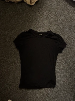Svart basic t-shirt från Gina Tricot - En enkel svart t-shirt från Gina Tricot i mjuk bomull. Klassisk rund halsringning och korta ärmar. Perfekt att matcha med jeans eller kjol för en clean och stilren look.