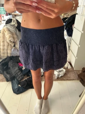 Mörkblå volangkjol med broderi - Kort mörkblå kjol med bred smock i midjan och volangig nederdel. Kjolen har broderade detaljer och hålmönster som ger en söt och trendig look. Perfekt för dig som gillar en feminin och avslappnad stil.
