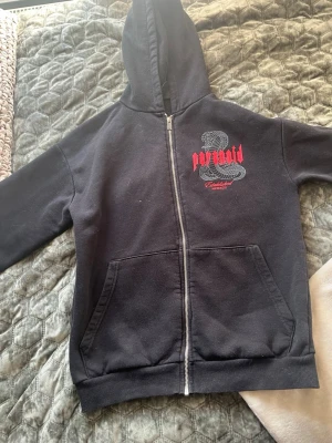 Svart Paranoid hoodie med ormtryck - Svart hoodie med dragkedja från Paranoid. Stor orm och röd text på både fram- och baksida, där det står 'paranoid' och 'Real eyes realize real lies'. Mjuk bomullskänsla, huva och två stora fickor framtill. Perfekt för dig som gillar streetwear och statement-plagg.