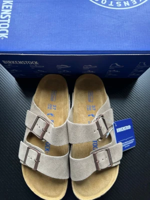 Beige Birkenstock Arizona sandaler - Säljer ett par klassiska Birkenstock Arizona sandaler i beige mocka med två breda remmar och justerbara metallspännen. Fotbädden är i mjukt naturfärgat material för extra komfort. Perfekta för en avslappnad och trendig stil.