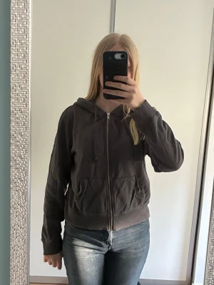Mörkgrå zip hoodie med huva - Säljer en mörkgrå hoodie med dragkedja och huva. Hoodien har två fickor framtill, ribbade muddar vid ärmslut och nederkant samt justerbara snören i huvan. Perfekt till jeans eller mjukisbyxor för en chill look.