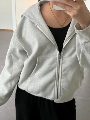 Ljusgrå zip hoodie med huva - Ljusgrå hoodie med dragkedja framtill och stor huva. Hoodien har två rymliga fickor på magen och ribbade muddar vid ärmslut och nederkant. Tillverkad i mjukt sweatshirtmaterial, perfekt för en chill och avslappnad stil.