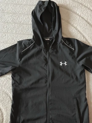 Svart hoodie från Under Armour - Snygg svart hoodie från Under Armour med dragkedja framtill och vit logga på bröstet. Hoodien har huva och diskreta vita detaljer vid axlarna. Perfekt för en sportig och avslappnad stil.
