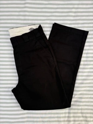 Dickies svart byxor, storlek W28 - Svarta byxor från Dickies. Dessa byxor har en klassisk passform med raka ben och hög midja. De har två sidfickor framtill och två passfickor baktill. Gylfen stängs med dragkedja och knapp.