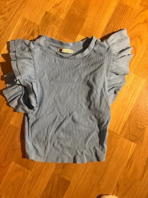 Blå volangtopp från Gina Tricot - Supersöt ljusblå topp från Gina Tricot med volanger på axlarna och ribbad struktur. Rund halsringning och korta ärmar med extra volym. Perfekt för en trendig och lekfull stil.