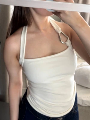 Vit asymmetrisk halterneck linnetopp - Trendig vit linnetopp med asymmetrisk halterneck och metallring på ena axeln. Toppen är ärmlös och har en tight passform som framhäver figuren. Perfekt för dig som vill sticka ut med en unik detalj i din outfit.