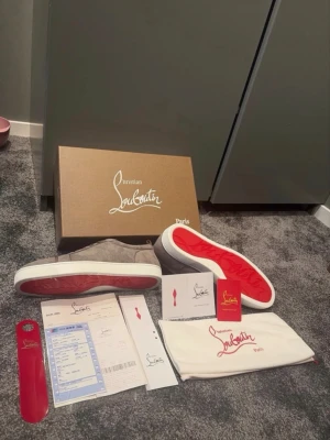 Gråa Christian Louboutin sneakers - Snygga gråa sneakers från Christian Louboutin med klassisk röd sula och vit kant. Skorna är låga och har en stilren design i mocka. Medföljer originalkartong, dustbag och tillbehör. Perfekt för dig som vill ha exklusiva skor med ikonisk look. Storlek 43! Vid frågor eller funderingar är de bara att höra av sig!