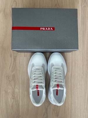 Vita sneakers från Prada - Snygga vita sneakers från Prada med meshpaneler och detaljer i läder. Skorna har en chunky sula och klassisk snörning. Röd logga på plösen ger en clean och exklusiv vibe. Perfekta för dig som vill ha en stilren och modern look.