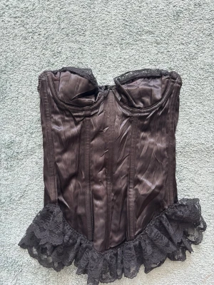 Svart satin korsett med spetsvolang - Snygg svart korsett i glansigt satinmaterial med vadderade kupor och dekorativ spetsvolang nedtill. Axelbandslös modell med markerade sömmar för en klassisk siluett. Perfekt för dig som vill ha en elegant och edgy look.