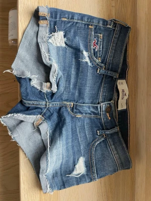 Blå jeansshorts från Hollister - Säljer ett par blå jeansshorts från Hollister med slitna detaljer och råa kanter. Shortsen har klassisk femficksdesign, låg midja och snygga slitningar framtill och baktill. 