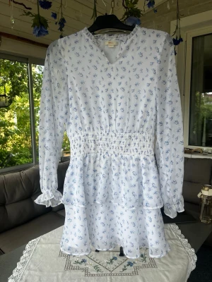 Vit blommig kortklänning med volang - Söt vit kortklänning med blått blommönster och volangdetaljer. Klänningen har långärmade ärmar med resår vid handlederna, smock i midjan och v-ringad hals. Perfekt för sommarens alla tillfällen. Den har en lite missfärgad fläck på axel (kolla bild) men inget som man tänker på 