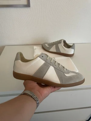 Maison margiela gats - Säljer ett par extremt stilrena och klassiska Maison Margiela Replica sneakers, även kända som German Army Trainers (GATs). Skorna är tillverkade i Italien av absolut högsta kvalitet i en kombination av mjukt lammskinn och kalvmocka. En tidlös modell i vitt och grått med den ikoniska gummisulan (gum sole) som passar till i stort sett allt. Info om skorna:  Märke: Maison Margiela Paris  Modell: Replica (German Army Trainers)  Storlek: 40  Material: Lammskinn och kalvmocka (Lambskin and calf-split