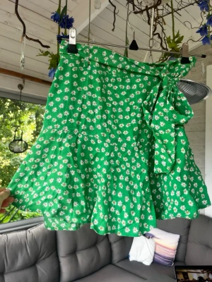 Grön blommig omlottkjol med volang - Superfin grön kortkjol med vita blommor och omlottmodell. Kjolen har en volangkant och knyts i midjan med ett brett band. Perfekt för vår och sommar och har innebyggda shorts och dragkedja på sidan 