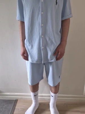Ljusblått set från Ralph Lauren - Säljer ett ljusblått set med Ralph Lauren. Kortärmad skjorta och matchande shorts. Setet är gjort i ett ribbat tyg och har vita knappar på skjortan samt den klassiska loggan broderad på bröstet och benet. Perfekt för en avslappnad och stilren look.
