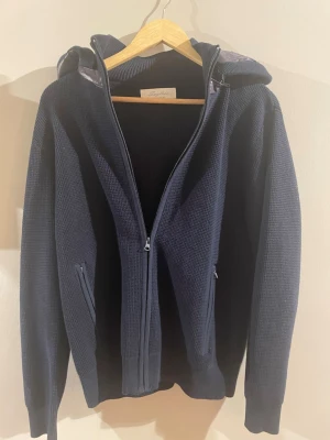 | Sayless | Mörkblå stickad cardigan  - Aldrig använd! Mörkblå stickad cardigan från Sayless med dragkedja framtill och två fickor. Huvan har ett quiltat, glansigt tyg i grått som ger en schysst kontrast. Perfekt för dig som gillar en avslappnad men ändå stilren look.