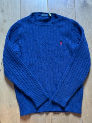 stickad tröja Polo Ralph Lauren - Mörkblå kabelstickad tröja från Polo Ralph Lauren med rund halsringning och röd broderad logga på bröstet. Tröjan har långa ärmar och ribbade muddar vid ärmslut och nederkant. Perfekt för dig som gillar klassisk och stilren design.