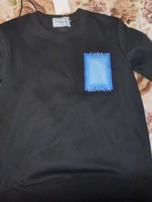 Svart sweatshirt från Casablanca - Svart sweatshirt från Casablanca med tryck i blått och vitt på bröstet och ryggen där det står 'Casa Blanca'. Tröjan har rund halsringning, ribbade muddar och är tillverkad i mjukt bomullsmaterial. Perfekt för dig som gillar stilrena plagg med grafiska detaljer.