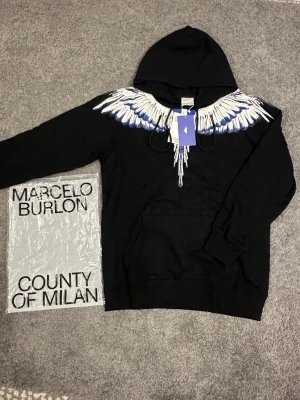 Svart hoodie Marcelo Burlon med vingar - Säljer en svart hoodie från Marcelo Burlon County of Milan med ikoniskt vingtryck i vitt och blått över axlar och bröst. Hoodien har huva med dragsko och en stor magficka. Perfekt för dig som gillar streetwear och vill sticka ut.