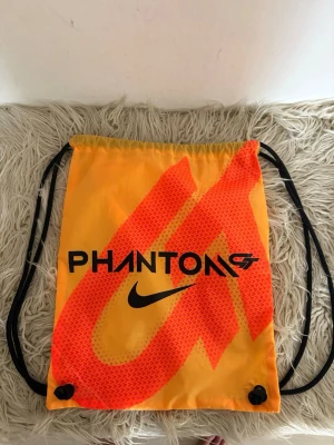 Nike Phantom GT gympapåse orange - Snygg orange gympapåse från Nike med svart text 'PHANTOM GT' och logga på framsidan. Väskan har svarta snören och är tillverkad i lätt syntetmaterial. Perfekt för träning eller att bära med sig det viktigaste. Cool design med röd grafik på framsidan.