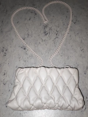 Vit handväska med kedjerem - Säljer en snygg vit handväska med quiltat mönster och kedjerem. Väskan har dragkedja upptill och är gjord i ett mjukt material som känns lyxigt. Perfekt för dig som vill ha en stilren accessoar med trendig känsla.