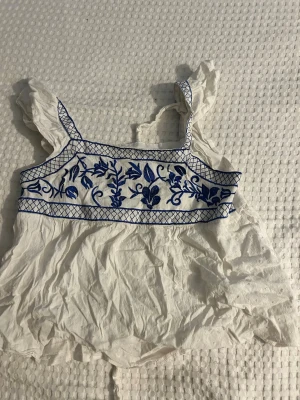 Vit och blå broderad linnetopp Shein - Supersöt vit linnetopp från Shein med blått broderi och volangdetaljer på axelbanden. Toppen har en luftig passform och är perfekt för varma dagar. Broderade blommor framtill ger en unik och trendig look.