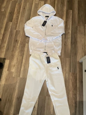 Vitt set från Polo Ralph Lauren - ALLA PRISFÖRSLAG ACCEPTERAS! Snyggt vitt set från Polo Ralph Lauren med zip-hoodie och matchande byxor. Setet har den klassiska loggan broderad på både tröjan och byxan. Tillverkat i mjukt material med bekväm passform och dragsko i midjan på byxorna. Perfekt för en avslappnad och stilren look.ALLA PRISFÖRSLAG ACCEPTERAS! Passar till M men också till S