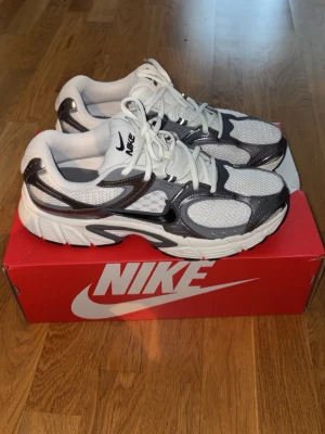 Nike vita och silverfärgade sneakers - Nike sneakers i vitt och silver med svarta detaljer. Skorna har en chunky sula, meshpaneler för bra ventilation och klassisk snörning. Swoosh-logga på sidan och bekväm vadderad insida. Perfekta för dig som gillar sportig och trendig stil.