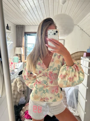 Blommig blus från Byic med puffärm - Supersöt blommig blus från Byic i ljus krämfärg med rosa, gröna och gula toner. Blusen har v-ringning, markerad midja och stora puffiga långärmar med resår vid ärmslut. Perfekt för en trendig och feminin look.