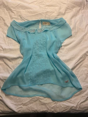 Ljusblå blus med pärldetaljer Hollister - Supersöt ljusblå blus från Hollister med korta ärmar och transparent, lätt material. Kragen är dekorerad med glittrande pärlor och blusen har en feminin, flowy passform. Perfekt för en fräsch och trendig look.