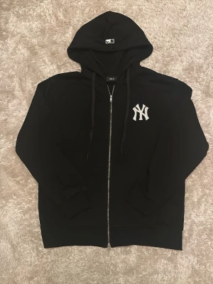 Svart Yankees zip hoodie med huva - Snygg svart hoodie med dragkedja från New Era, prydd med det klassiska New York Yankees-loggan i vitt på bröstet. Hoodien har justerbar huva med snören och ribbade muddar. Perfekt för dig som gillar sportig streetstyle.