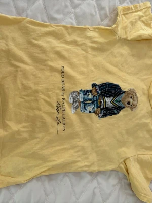 Gul Polo Bear t-shirt från Ralph Lauren - Snygg gul t-shirt från Ralph Lauren med ikoniska Polo Bear-trycket framtill. T-shirten har rund hals, korta ärmar och är gjord i mjuk bomull. Perfekt för dig som gillar streetstyle och vill ha något unikt i garderoben.