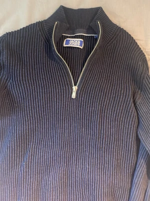 Mörkblå ribbstickad tröja Jack & Jones - Mörkblå ribbstickad tröja från Jack & Jones med halv dragkedja och hög krage. Tröjan har en cool struktur med breda ribbor och är perfekt för lager på lager. Klassisk och enkel design som funkar till det mesta. Storlek 164 (herr S/XS)