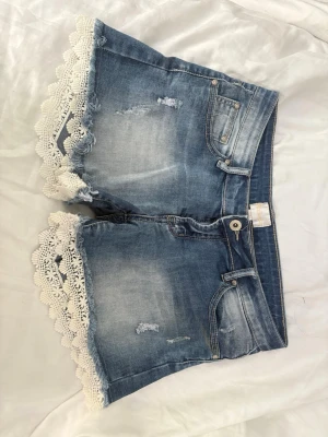 Lågmidjade Blå jeansshorts med spetskant - Säljer ett par blå jeansshorts med snyggt slitna detaljer och vit spetskant längst ner. Shortsen har klassisk femficksdesign, knapp och dragkedja framtill. Perfekta för sommaren och ger en söt och trendig look.