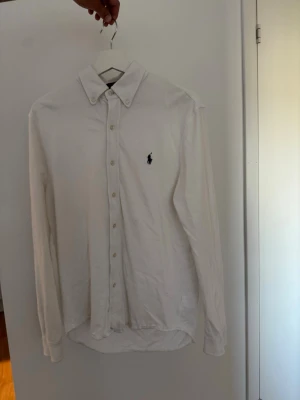 Vit skjorta från Ralph Lauren - Klassisk vit skjorta från Ralph Lauren med button-down krage och broderad logga på bröstet. Tillverkad i lätt bomullsmaterial (featherweight mesh) och har långa ärmar samt knappar framtill. Perfekt till en clean och stilren look.