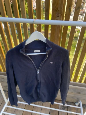 Mörkblå ziptröja från Gant - Mörkblå ziptröja från Gant med liten broderad logga på bröstet. Tröjan har hög krage, dragkedja framtill och ribbade muddar vid ärmslut och nederkant. Perfekt för en avslappnad och stilren look. Storleken är 164 barn och den är i helt nytt skick. Nypris minst 500 kr
