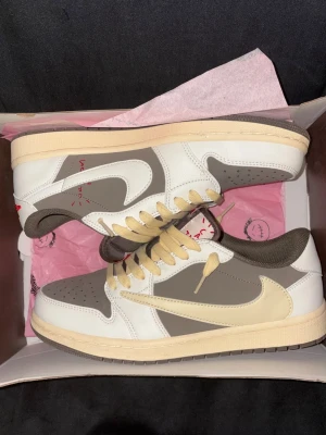 Nike x Travis Scott grå/beige sneakers - Nike x Travis Scott sneakers i low modell med beige, grå och vita paneler. Skorna har den ikoniska omvända swoosh-loggan på sidan, beige mellansula och grå yttersula. Ovandelen är i läder och mocka med beige skosnören och mörkbrunt foder. Perfekt för dig som gillar unik streetwear.