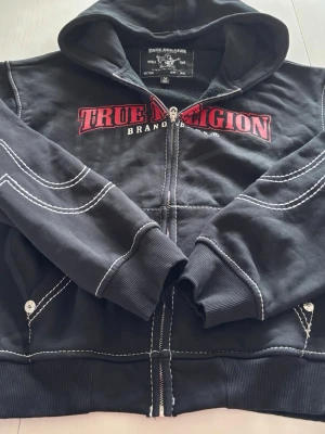 Svart True Religion hoodie aldrig använd kostar 1599 ny - Svart hoodie från True Religion med vit kontrastsöm och röd logga på bröstet. Dragkedja framtill, två fickor med knappdetaljer och stor huva. Mjuk insida och ribbade muddar vid ärmar och nederkant. Perfekt för en avslappnad streetwear-look.