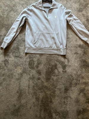 Ljusgrå quarter zip hoodie Polo Ralph Lauren - Ljusgrå hoodie från Polo Ralph Lauren med quarter zip och klassisk krage. Har en stor magficka och det ikoniska broderade logot på bröstet. Tillverkad i mjukt bomullsmaterial och har ribbade muddar vid ärmar och nederkant. Perfekt för en chill och stilren look.