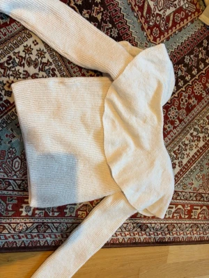 Beige stickad tröja med volang - Säljer en beige stickad tröja med stor volangdetalj över axlarna. Tröjan har långa ärmar och är ribbstickad för en snygg och bekväm passform. Perfekt för dig som gillar trendiga detaljer och en mjuk känsla.