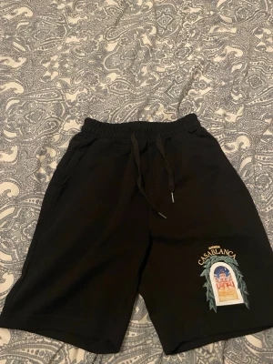 Svarta shorts från Casablanca - Svarta shorts från Casablanca med broderad färgglad logga och motiv på ena benet. De har elastisk midja med snörning och två sidofickor. Perfekta för en chill och stilren look.