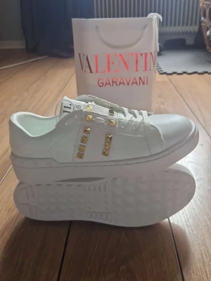 Valentino Rockstud vita sneakers - Snygga vita sneakers från Valentino Garavani med guldiga nitar på sidorna och breda band över ovandelen. Skorna är låga och har en chunky sula. Tillverkade i läder med klassisk snörning och coola detaljer som sticker ut.